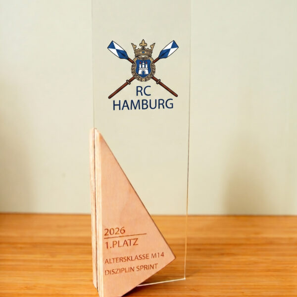 Ein hoher, rechteckiger Pokal, der auf einem Sockel steht und aus zwei verschiedenen Materialien besteht: Holz und Acryl. Der Großteil des Pokals ist eine matte Glasplatte, die oben und unten von passenden, winkelförmigen Holzstücken eingefasst ist. Das Holz hat eine helle, natürliche Maserung und Form eines Dreiecks, das sich von der Mitte nach außen zieht. In der Mitte der Glasplatte befindet sich ein gedrucktes Wappen. Der untere Holzsockel ist mit graviertem Text versehen: "2026", "2. PLATZ"